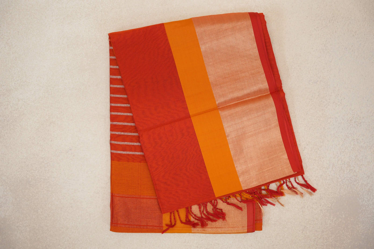 Folded tomato red Maheshwari silk cotton saree with a mustard orange stripe, pale border panel, red selvedge and tassels; striped pallu peeks out beneath.