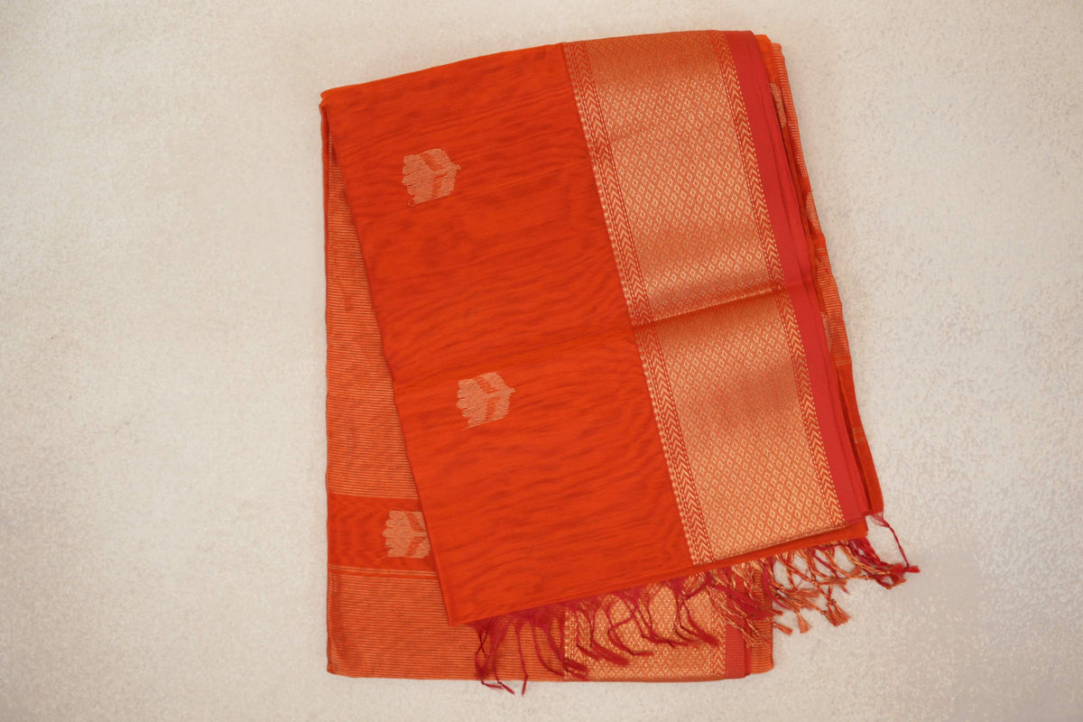Folded reddish orange Maheshwari silk cotton saree with small gold motifs, a wide geometric gold border, red selvedge and tassels.