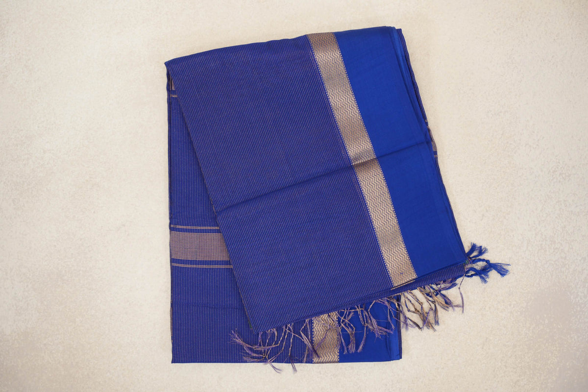 Folded deep royal blue Maheshwari silk-cotton saree with fine stripes, a gold-toned zigzag border, a plain blue edge and tassels.