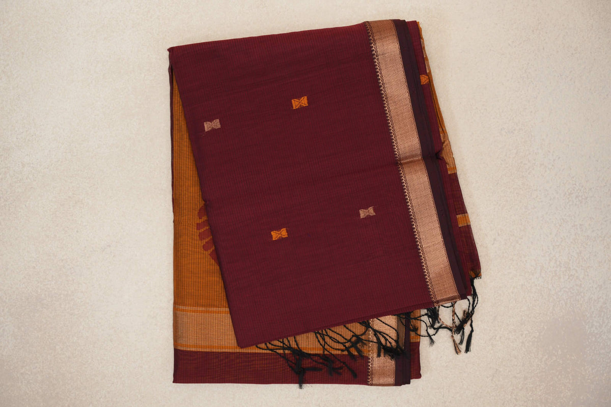 Folded deep maroon Maheshwari silk cotton saree with small orange and beige accents, a copper-toned patterned border, mustard orange fabric and dark tassels.