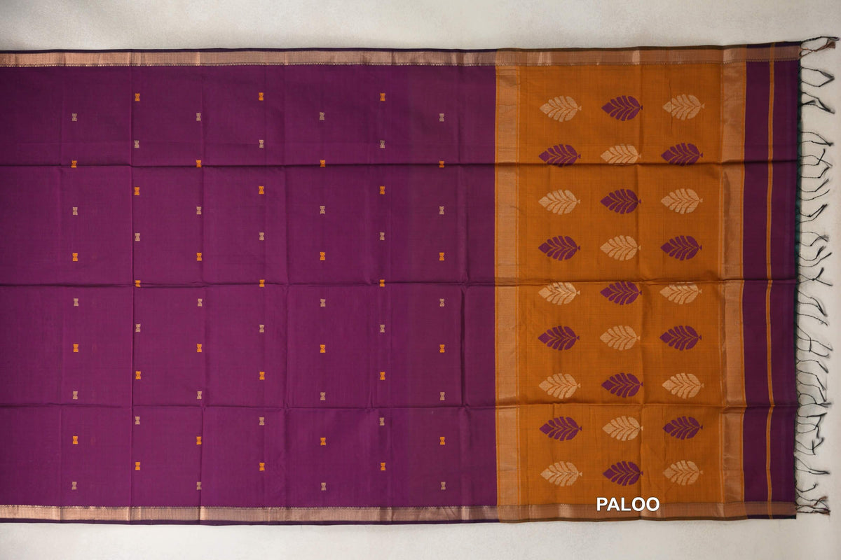 Maheshwari silk cotton saree laid flat, with a deep magenta body dotted with small motifs, gold-toned borders and a deep mustard orange pallu with leaf motifs and tassels.