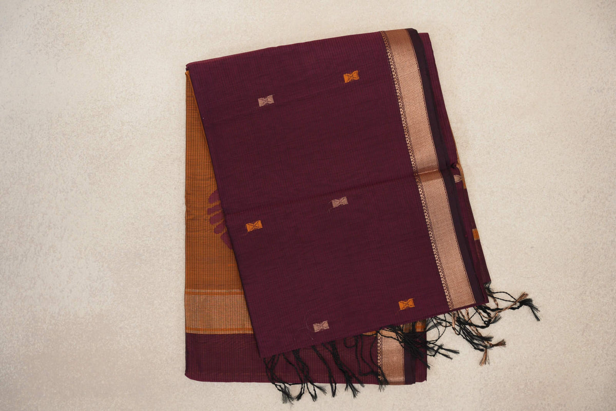 Folded deep wine Maheshwari silk cotton saree with small orange and beige motifs, a patterned border, mustard orange fabric beneath and black tassels.