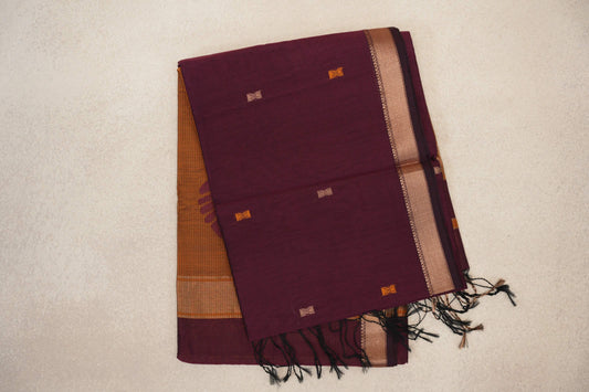 Folded deep wine Maheshwari silk cotton saree with small orange and beige motifs, a patterned border, mustard orange fabric beneath and black tassels.