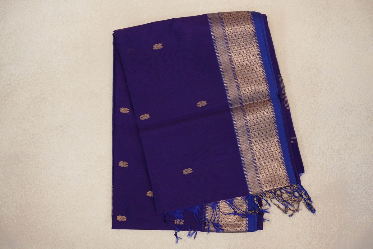 Folded royal purple Maheshwari silk cotton saree with small gold motifs, a wide geometric border, blue edging and tassels.
