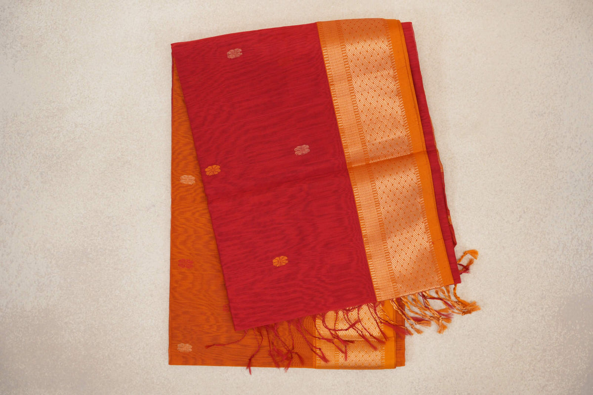 Folded tomato red Maheshwari silk cotton saree with small scattered motifs, a patterned mustard orange border with red selvedge, and loose tassels.