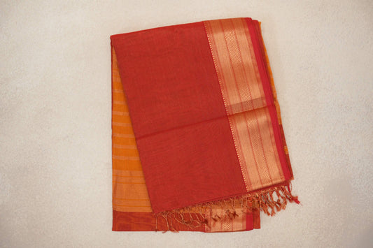 Folded tomato red Maheshwari silk cotton saree with fine stripes, a patterned gold-toned border, tassels and deep orange fabric peeking beneath.