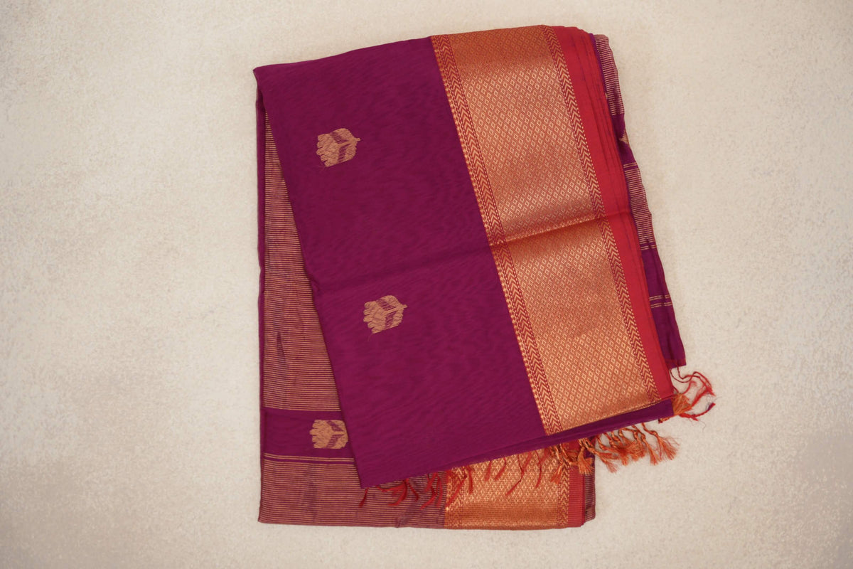 Folded reddish magenta Maheshwari silk cotton saree with small gold-toned motifs, a patterned border, pinkish red selvedge and tassels.