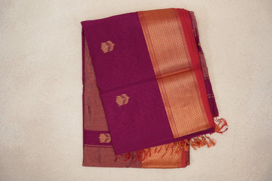 Folded reddish magenta Maheshwari silk cotton saree with small gold-toned motifs, a patterned border, pinkish red selvedge and tassels.