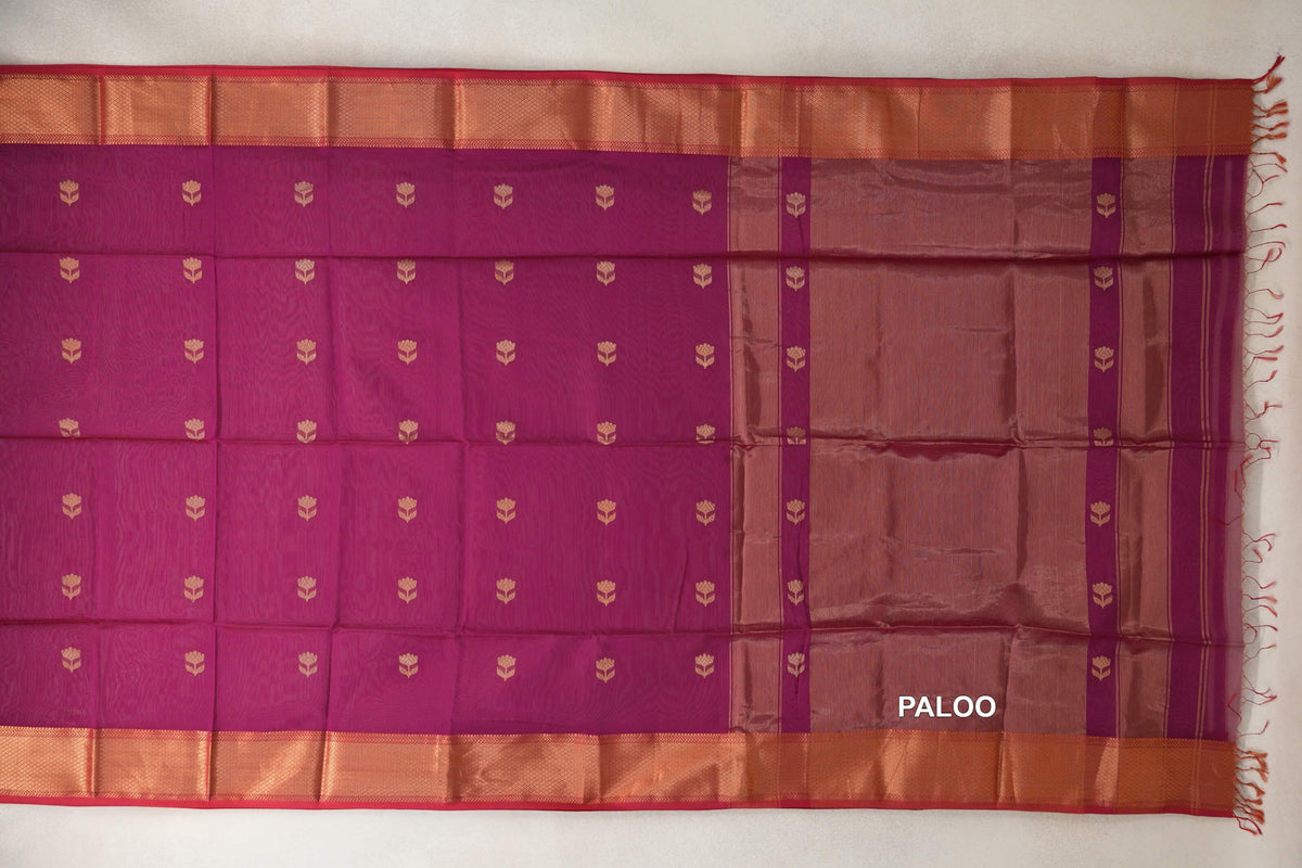 Reddish magenta Maheshwari silk cotton saree laid flat, with gold-toned floral motifs, copper-toned borders, pinkish red edges and a striped pallu with tassels.