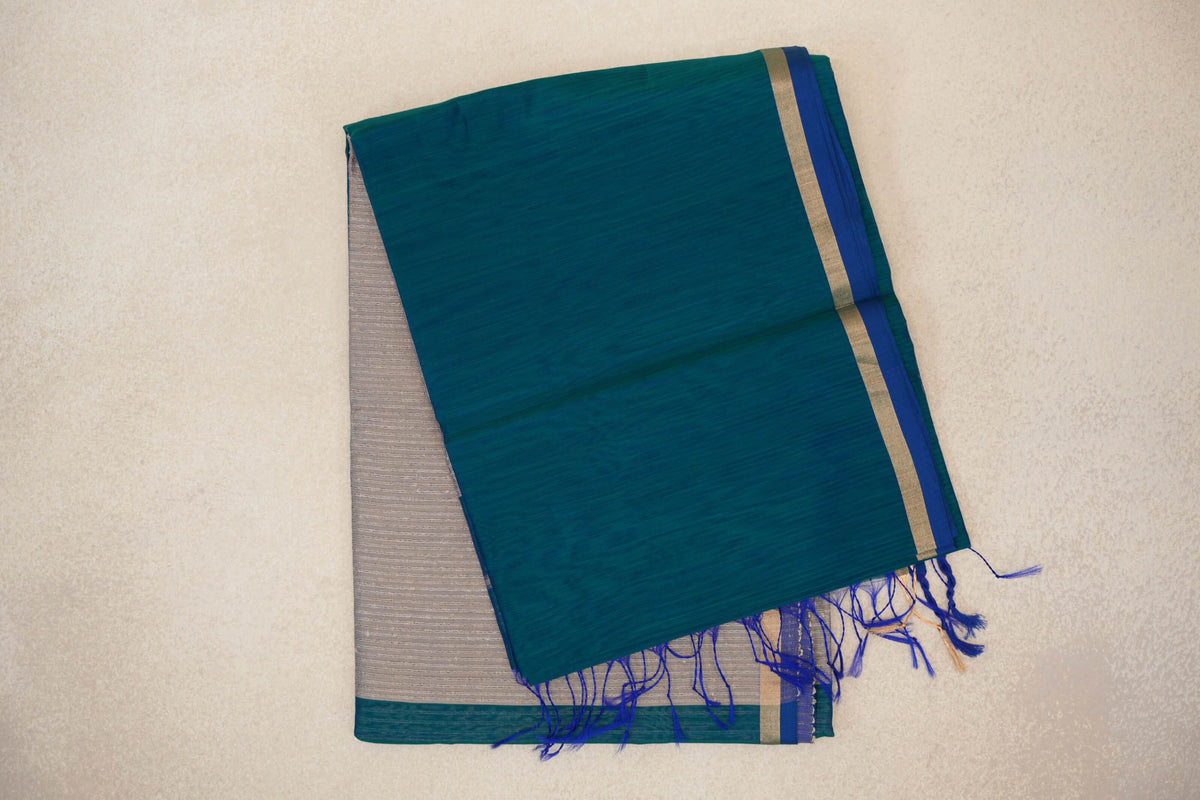 Folded Maheshwari silk cotton saree with a peacock green body, peacock blue selvedge border, and a silver beige pallu peeking out beneath blue tassels.