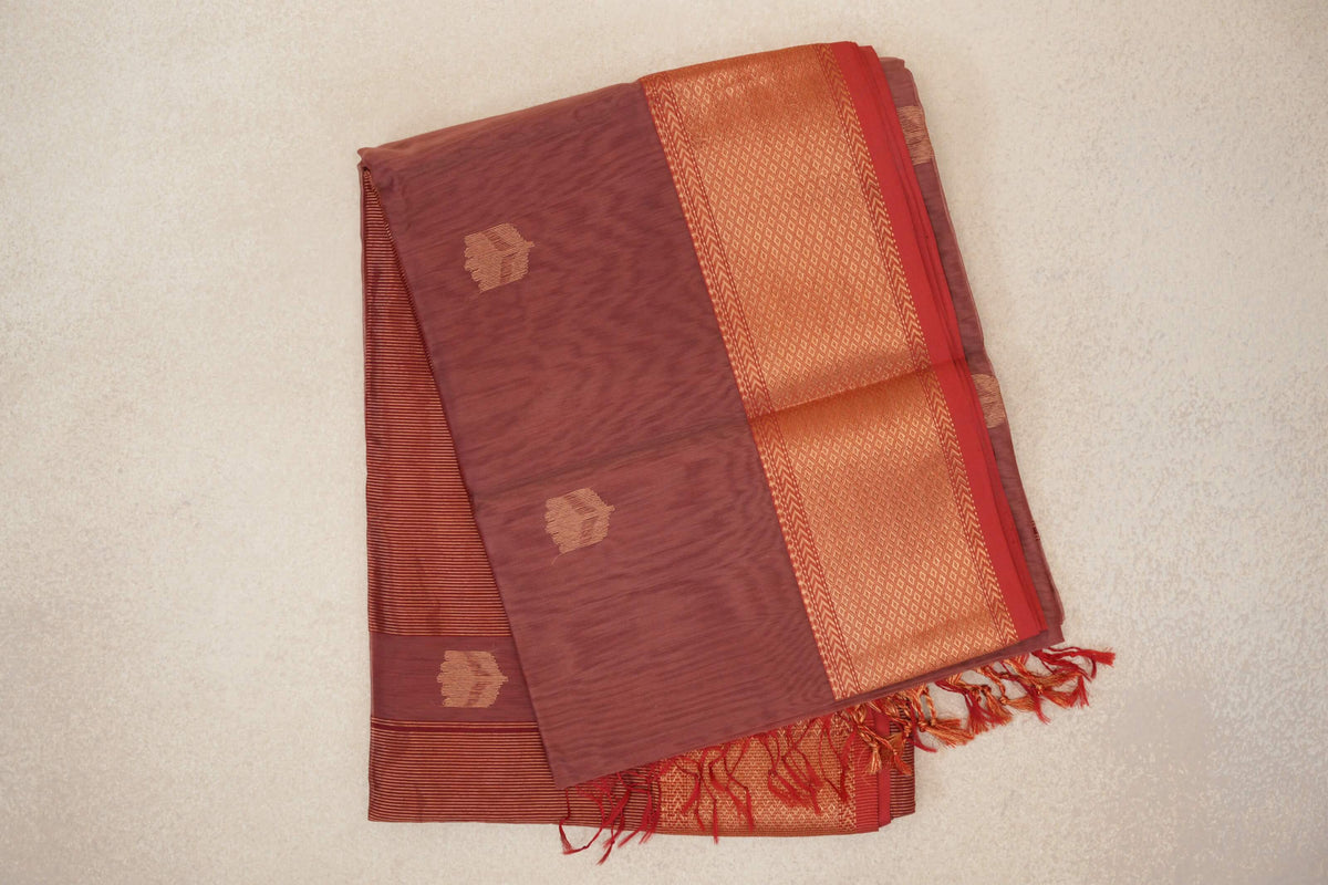 Folded dusty rose Maheshwari silk cotton saree with small gold-toned motifs, a red-edged geometric border and a fringed end.