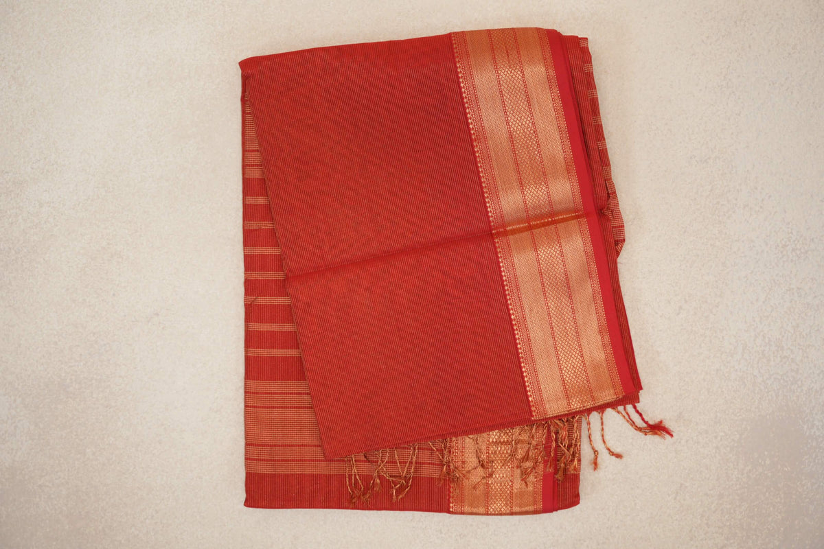 Folded tomato red Maheshwari silk cotton saree with fine gold stripes, a wide patterned gold border and tassels at one end.