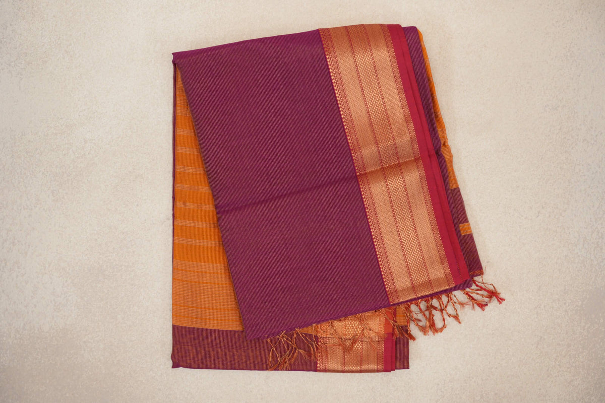Folded deep magenta silk cotton saree with fine stripes, a patterned border with rani pink selvedge, and deep orange fabric and tassels peeking out.
