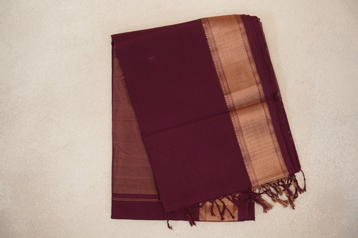 Folded deep wine Maheshwari silk-cotton saree with a broad gold-toned woven border, fine stripes on the underfold and tassels.