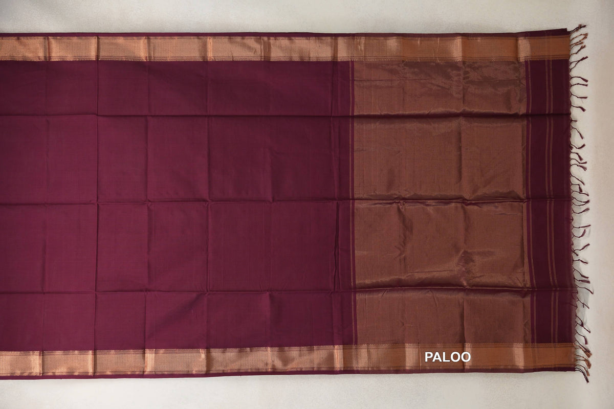 Deep wine Maheshwari silk-cotton saree laid flat, with a plain body, copper-toned borders and a broad pallu with tassels.