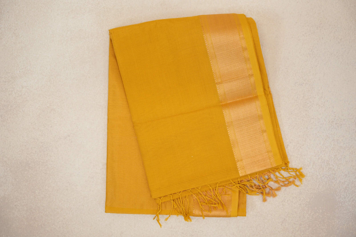 Folded turmeric yellow Maheshwari silk cotton saree with a fine checked weave, a shimmering woven border and tassels at one end.