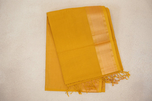 Folded turmeric yellow Maheshwari silk cotton saree with a fine checked weave, a shimmering woven border and tassels at one end.