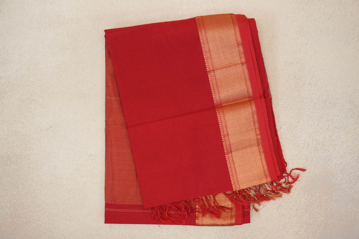 Folded vermilion red Maheshwari silk cotton saree with a broad gold-patterned border, fine stripes on the inner fold and red tassels.