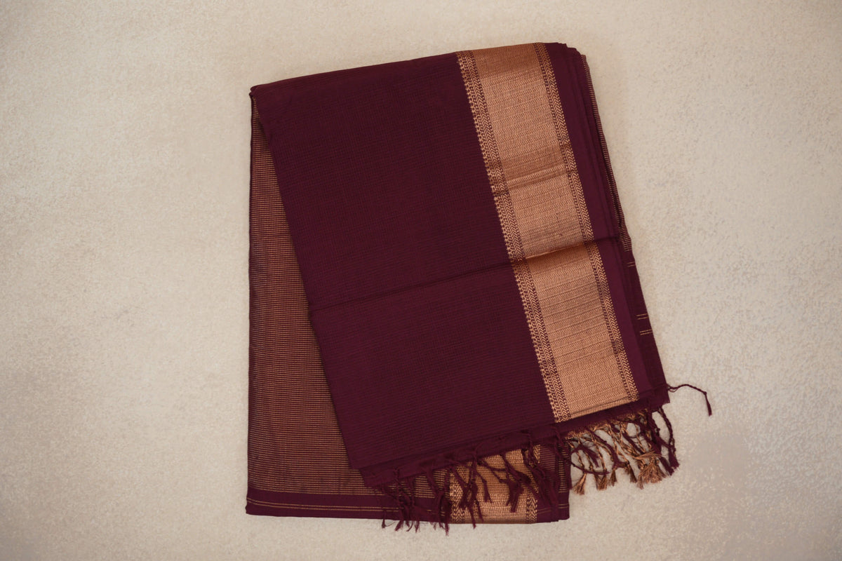 Folded deep wine Maheshwari silk cotton saree with a broad copper-gold woven border, fine stripes and tassels at one end.