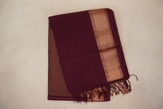 Folded deep wine Maheshwari silk cotton saree with a broad copper-gold woven border, fine stripes and tassels at one end.
