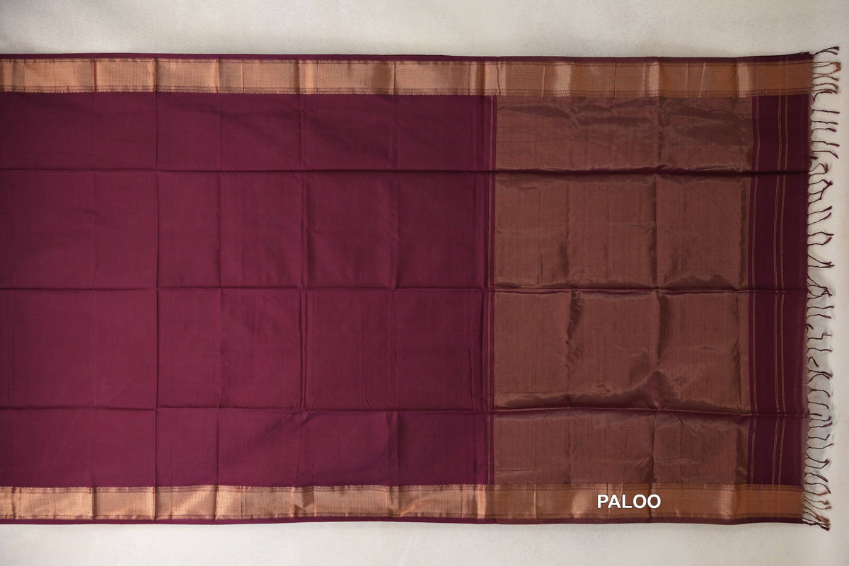 Deep wine Maheshwari silk cotton saree laid flat, showing a plain body, copper-toned borders, a shimmering pallu and end tassels.