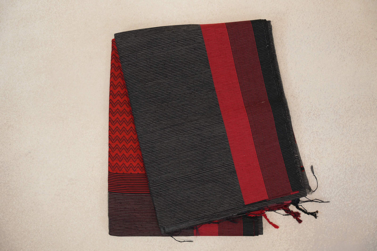 Folded charcoal grey Maheshwari silk cotton saree with fine stripes, a red and black border, red zigzag fabric peeking beneath and loose tassels.