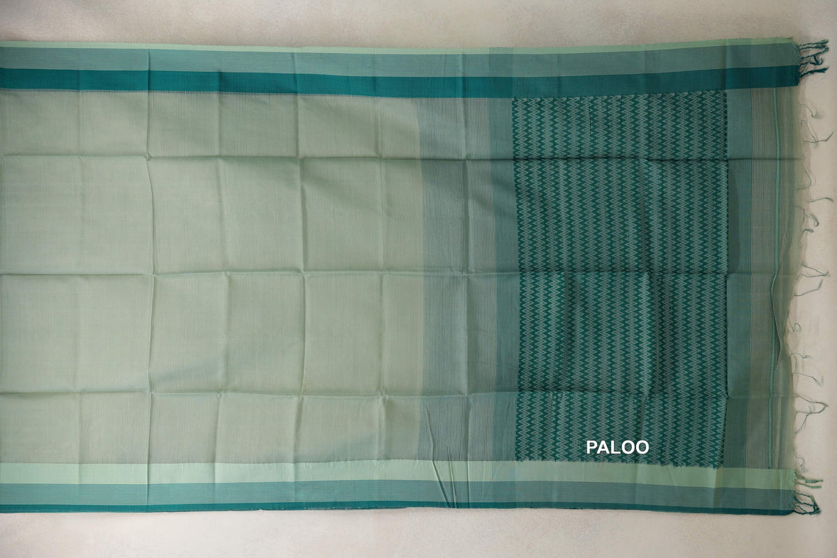 Maheshwari silk cotton saree spread out, with a pale sage green body, light sage borders edged in deep teal, and a zigzag-patterned pallu with tassels.