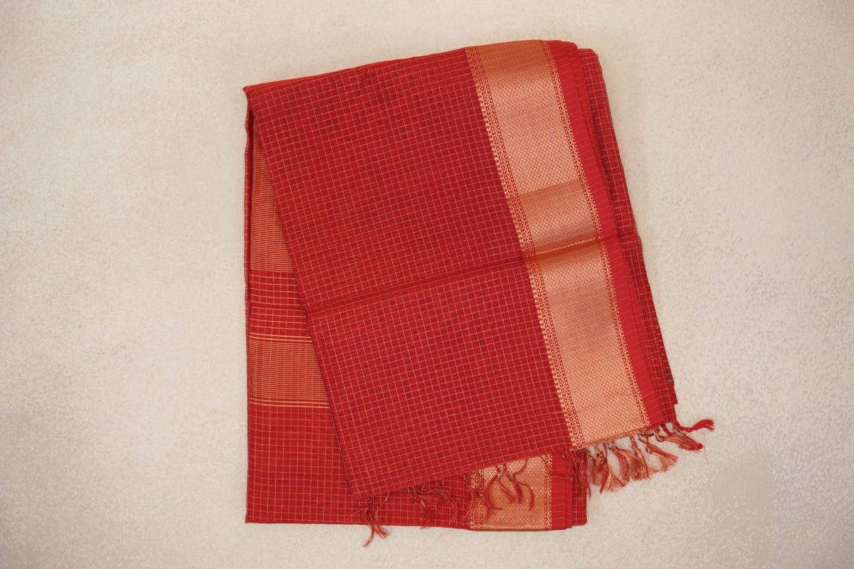Folded tomato red Maheshwari silk cotton saree with fine gold checks, a broad gold zari border and red tassels at the end.