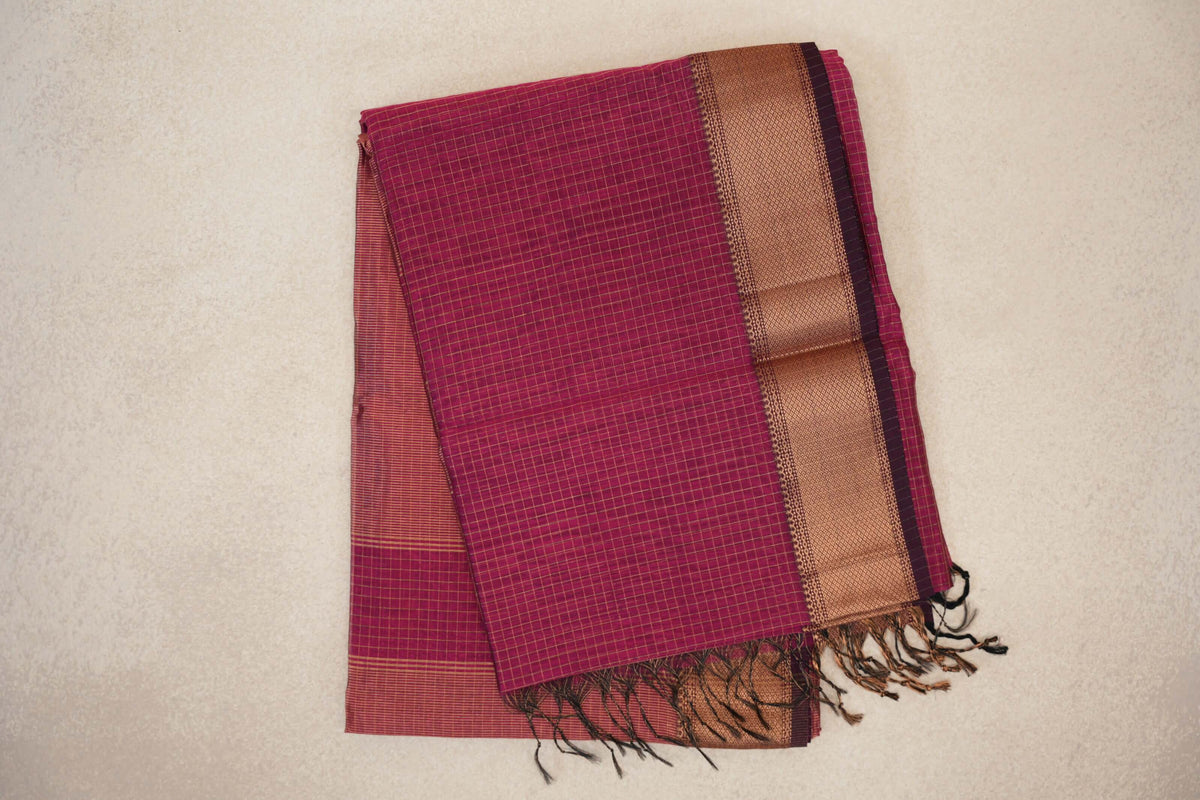 Folded Rani Pink Maheshwari silk cotton saree with a fine checked pattern, a broad copper-toned woven border, deep pink selvedge and tasselled ends.
