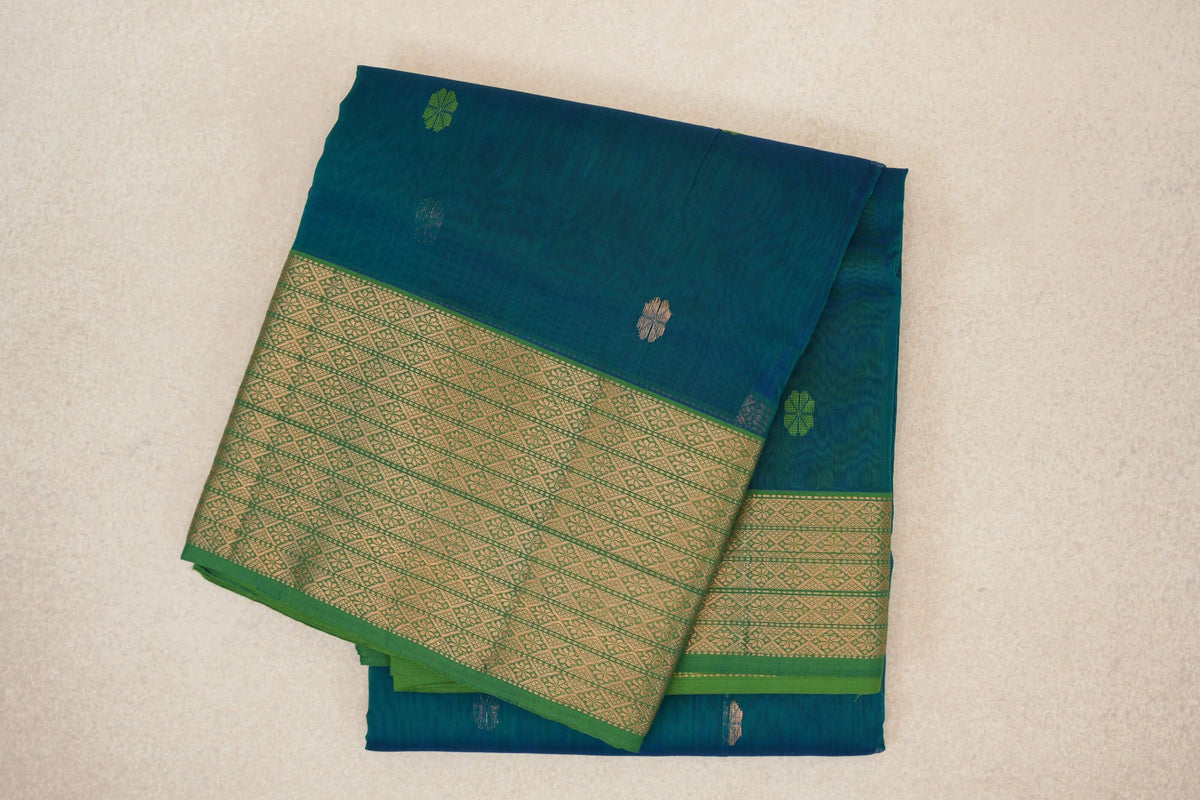 Folded teal blue Maheshwari silk cotton saree with small floral motifs and a leaf green border woven with gold-toned geometric patterns.