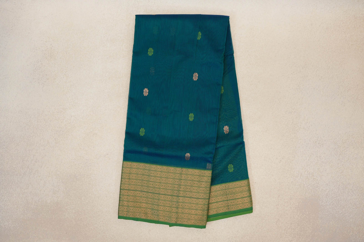 Folded teal blue Maheshwari silk cotton saree with scattered small motifs and a leaf green border woven with gold geometric patterns.