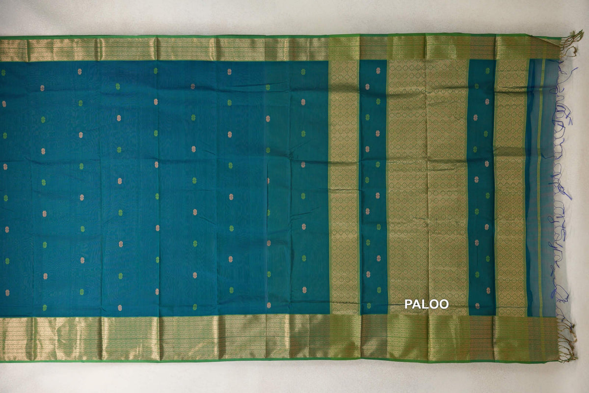 Maheshwari silk cotton saree spread flat, with a teal blue body dotted with small motifs, leaf green patterned borders and a geometric, fringed pallu.