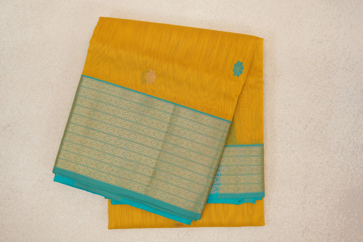 Folded mango yellow Maheshwari silk cotton saree with small floral motifs and a wide turquoise blue border woven with geometric patterns.