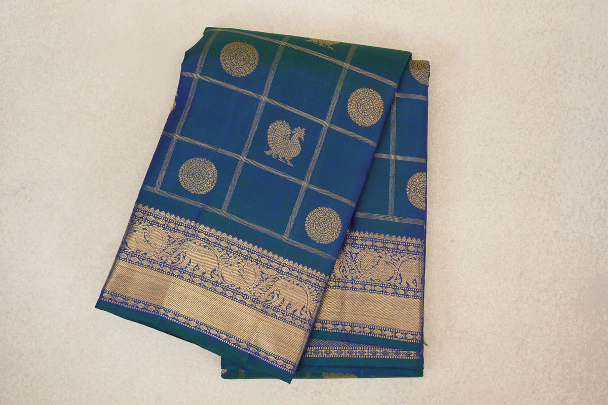 Folded peacock blue Kanjivaram silk saree with a gold grid, circular medallions, a peacock motif and a wide elephant-patterned border.