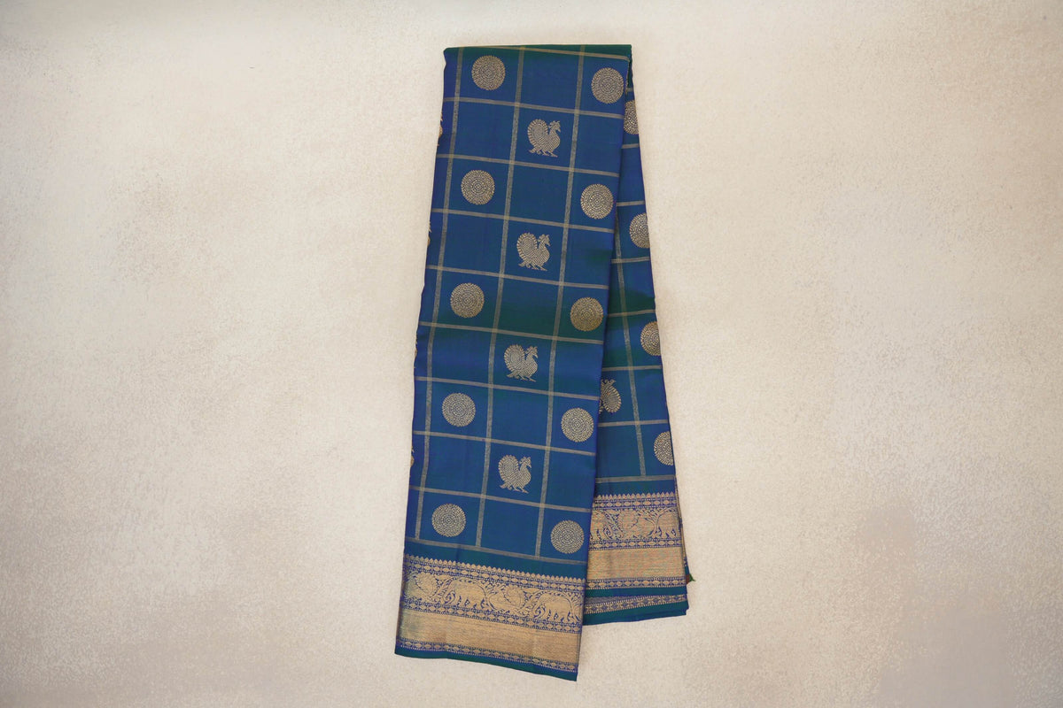 Folded peacock blue Kanjivaram silk saree with a gold grid, circular and peacock motifs, and an ornate elephant border.