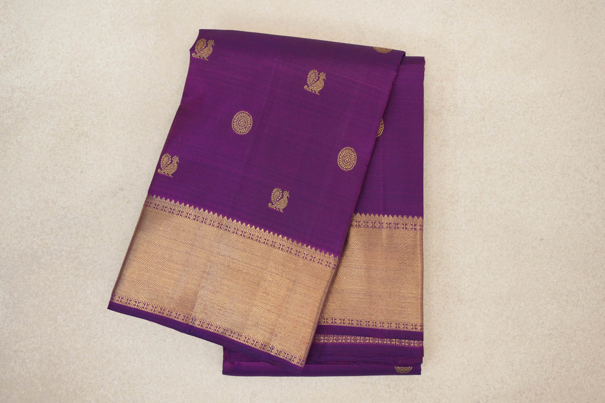 Folded purple Kanjivaram silk saree with gold peacock and circular motifs and a broad gold border with geometric trim.