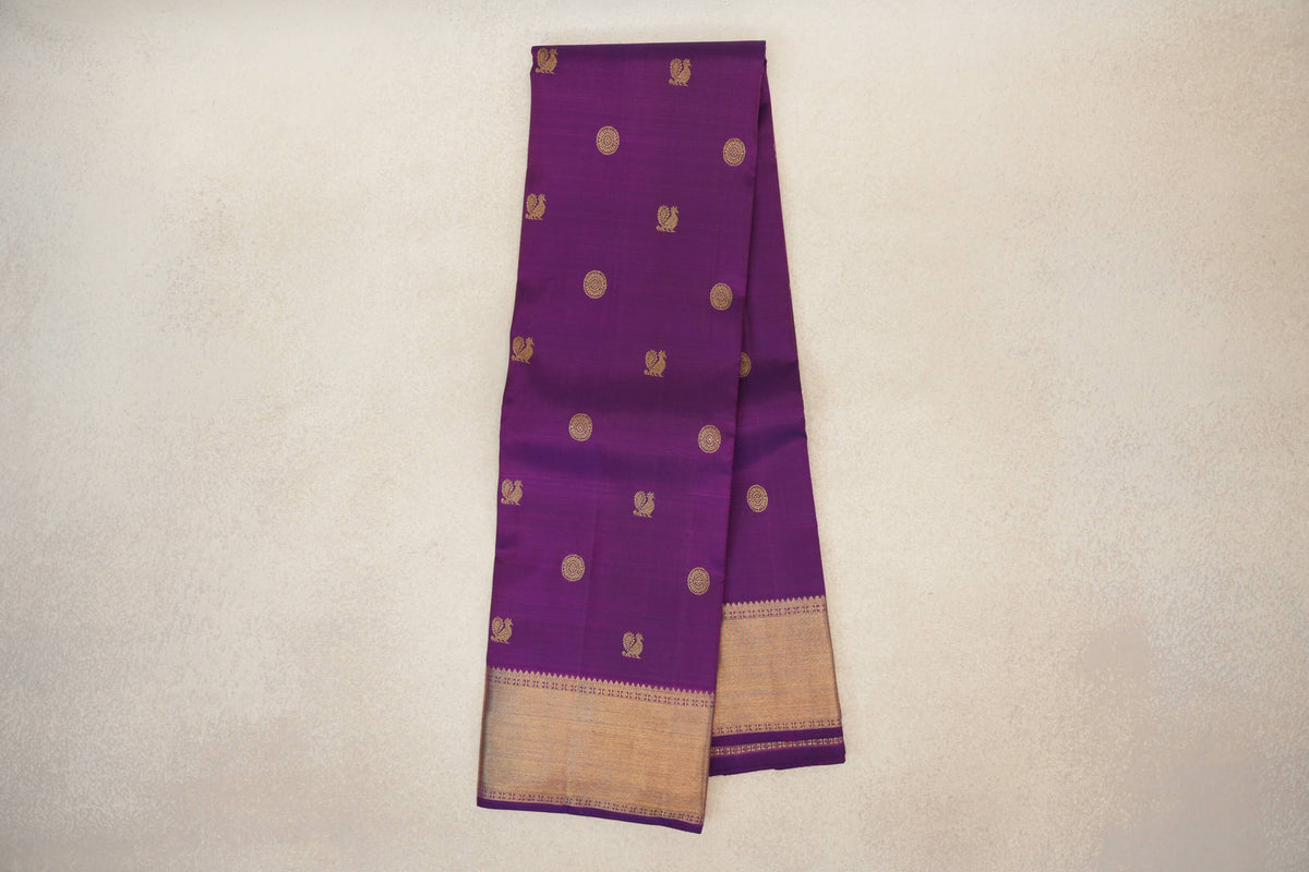 Folded purple Kanjivaram silk saree with scattered gold bird and circular motifs and a broad gold-toned border at the bottom.