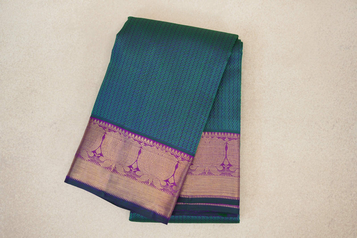 Folded peacock green Kanjivaram silk saree with a fine zigzag pattern and a purple border featuring gold-toned woven designs and bottle green edging.
