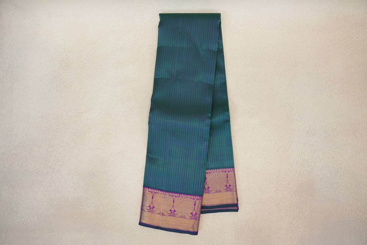Folded peacock green Kanjivaram silk saree with a fine zigzag pattern and purple borders featuring gold-toned lotus motifs and bottle green edging.