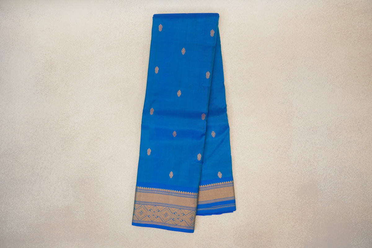 Folded peacock blue lightweight Kanchipuram silk saree with scattered gold motifs and a wide patterned gold border.
