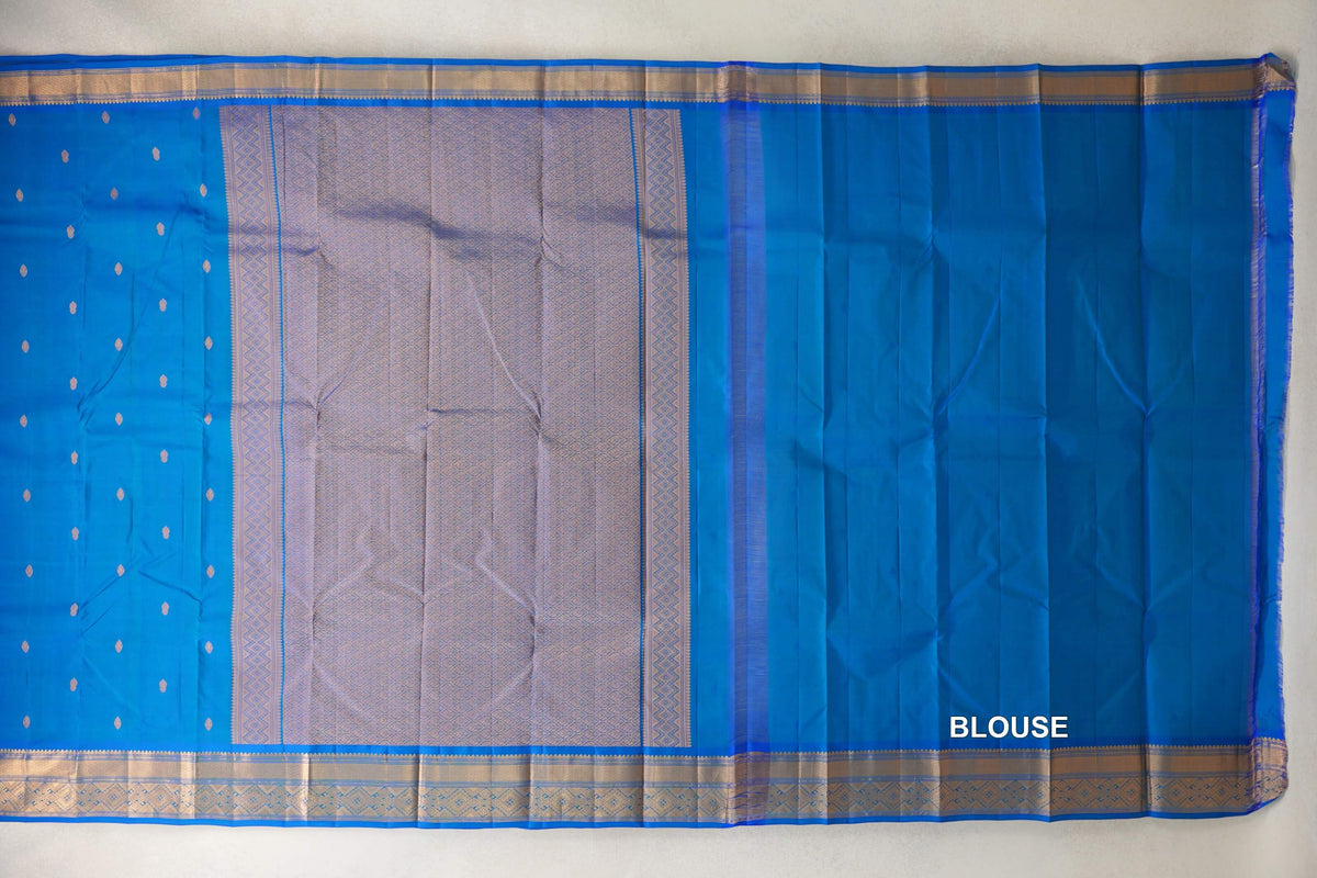 Peacock blue lightweight Kanchipuram silk saree laid flat, showing a body with small gold motifs, a patterned zari pallu and borders, and blouse fabric at right.