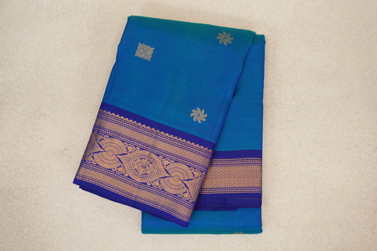 Folded peacock blue Kanchipuram silk saree with scattered gold floral motifs and a deep royal blue border woven with ornate gold zari patterns.