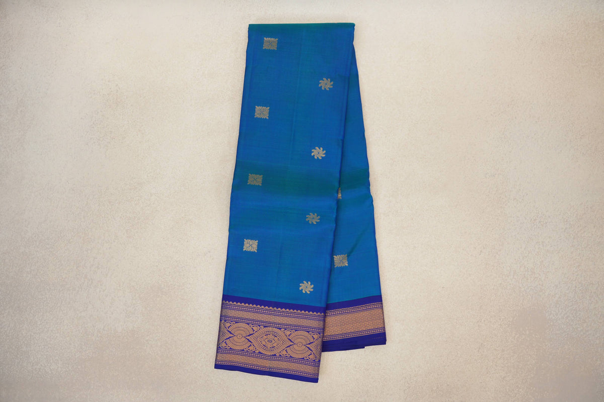 Folded peacock blue Kanchipuram silk saree with scattered gold motifs and a deep royal blue border woven with ornate gold designs.