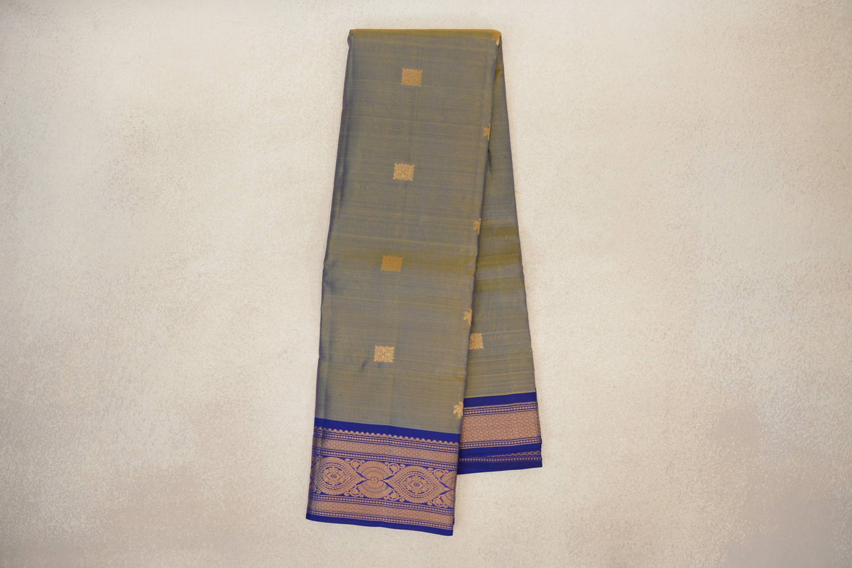 Folded Kanchipuram silk saree showing a muted yellow and violet body with small gold square motifs and a deep royal blue border with gold patterns.
