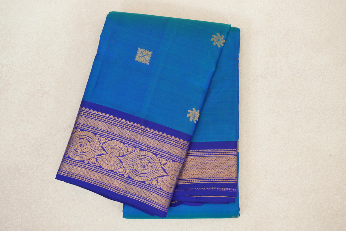 Folded lightweight Kanchipuram silk saree with a peacock blue body, small gold floral motifs and a deep blue violet border with ornate gold patterns.