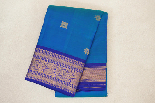 Folded lightweight Kanchipuram silk saree with a peacock blue body, small gold floral motifs and a deep blue violet border with ornate gold patterns.