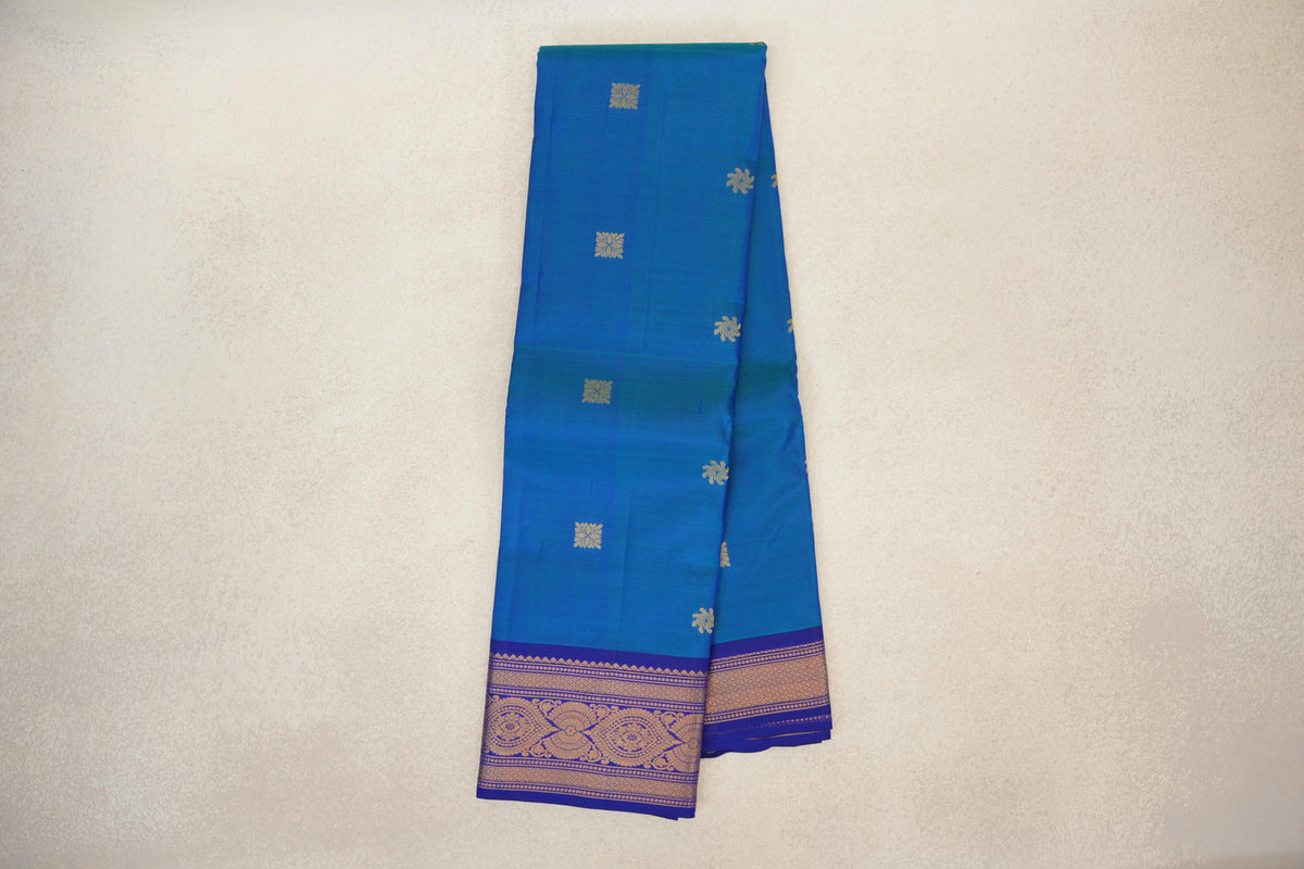 Folded peacock blue lightweight Kanchipuram silk saree with scattered gold floral motifs and a deep blue violet border with gold paisley and floral designs.