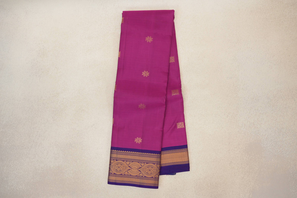 Folded deep magenta Kanchipuram silk saree with scattered gold floral motifs and violet purple borders woven with gold patterns.