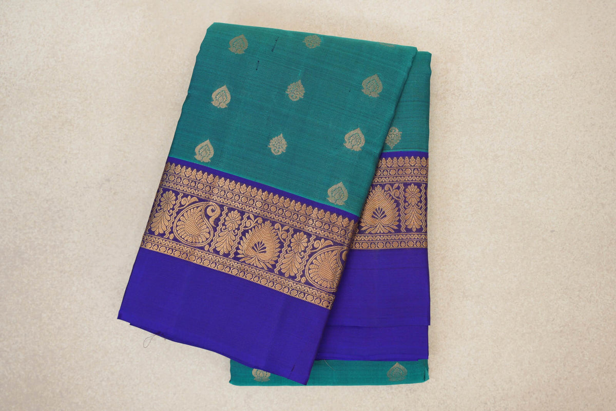 Folded lightweight Kanchipuram silk saree with a peacock green body, small gold paisley motifs and a deep royal blue border with gold floral designs.