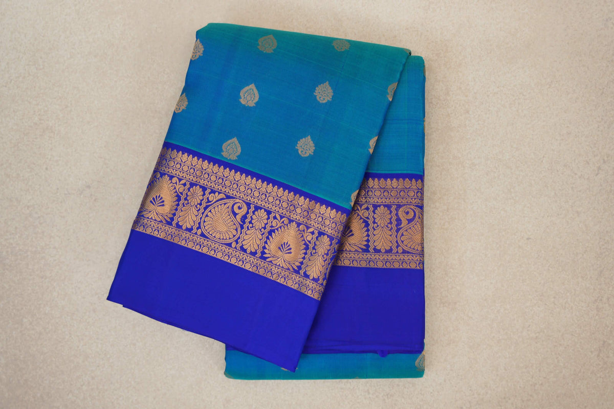 Folded peacock blue Kanchipuram silk saree with small gold motifs and a royal blue border patterned with gold paisleys and flowers.